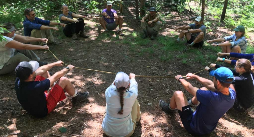 a group of people sit in a circle and hold a rope as part of a teamwork exercise. 
